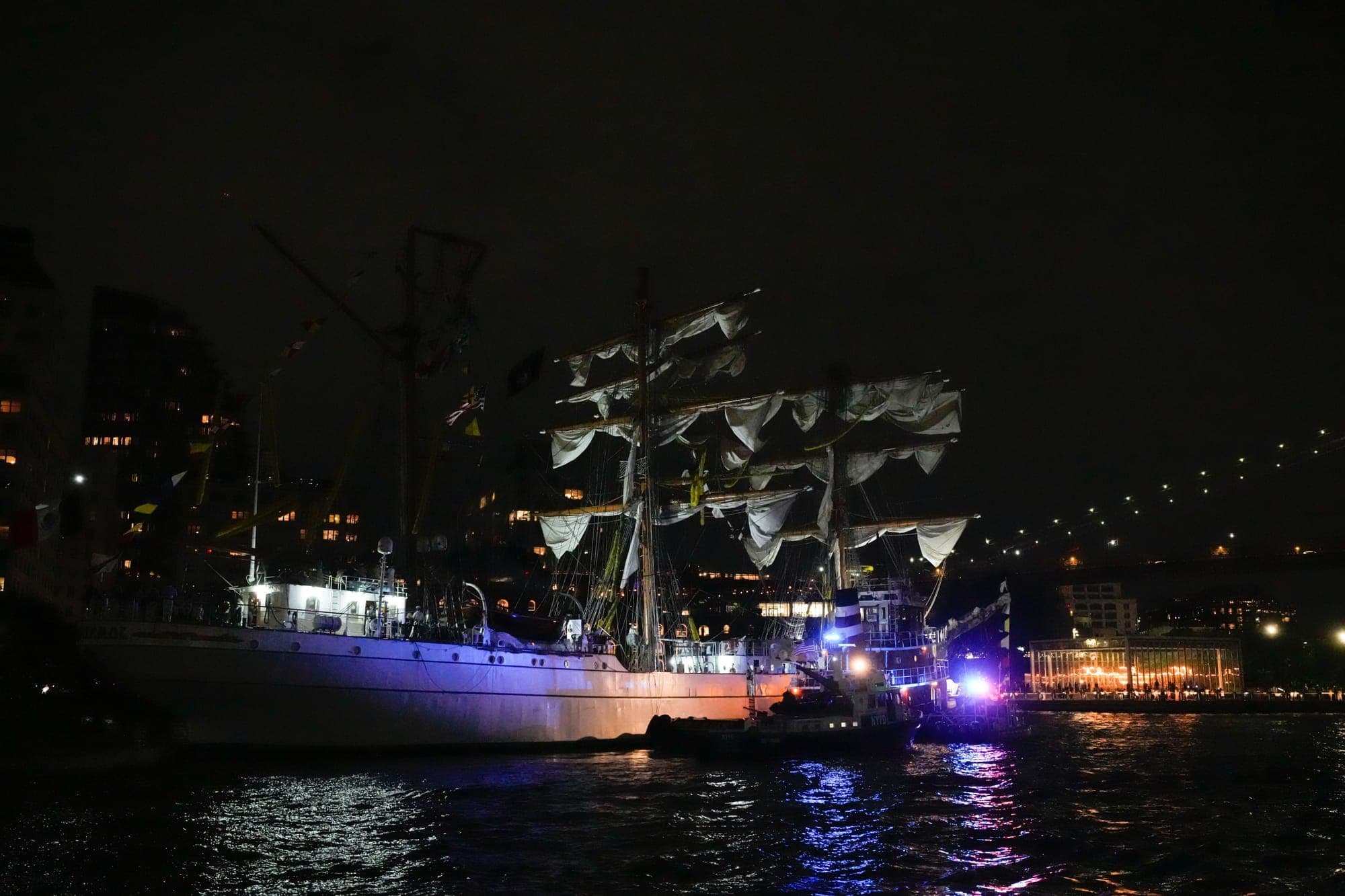 A nighttime scene shows a tall ship docked near the Brooklyn Bridge, illuminated by city lights and emergency boats with flashing blue and red lights reflecting on the water.