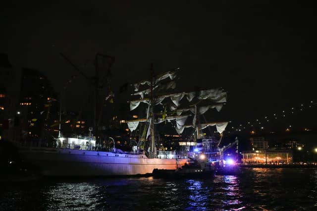 A nighttime scene shows a tall ship docked near the Brooklyn Bridge, illuminated by city lights and emergency boats with flashing blue and red lights reflecting on the water.