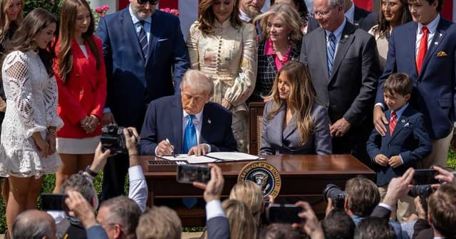 President Trump signs the Take It Down Act at the White House, joined by Melania Trump, officials, and supporters, in front of U.S. flags during the signing ceremony.